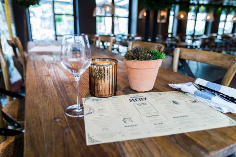 Träbord med vinglas, bestick, tygservett, liggande meny, blomkruka och ljuslykta. Restaurangsalen suddig i bakgrunden.