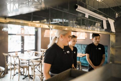 Servitörer som står vid kockarnas bänk med restaurangens bord och stolar i bakgrunden.