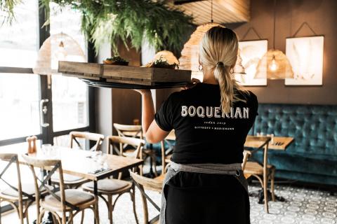Ryggtavlan på en servitör som bär en stor bricka med tapaskorgar över vänster axel. Texten Boquerian stort ryggen och restaurangsalen i bakgrunden.